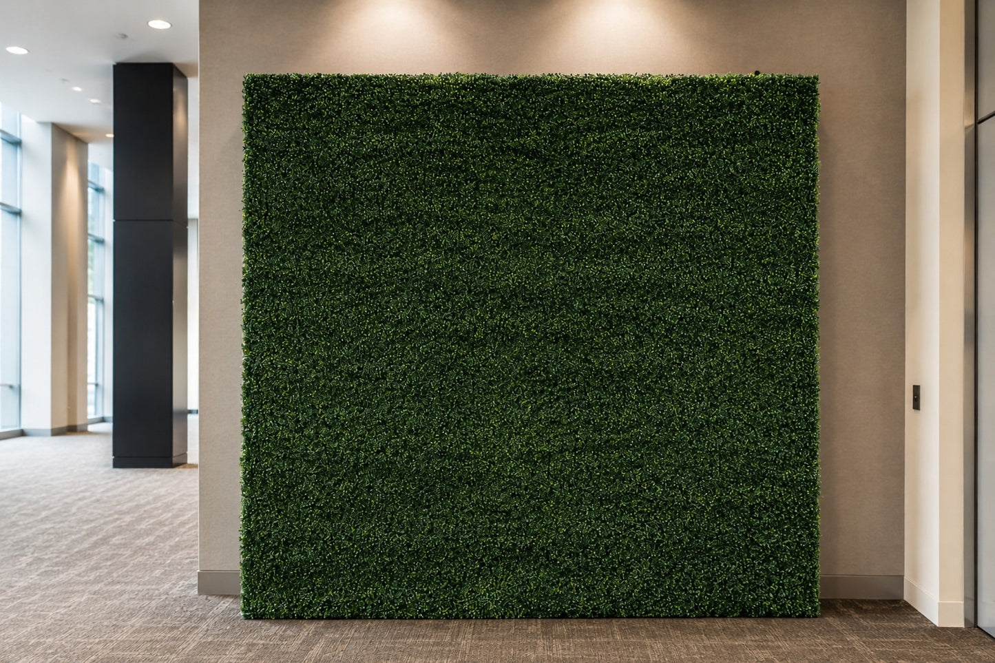 A large rectangular artificial green hedge wall mounted on a beige wall in an indoor office or hallway space with carpeted floor and recessed lighting.