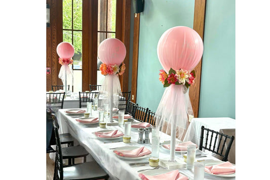 Decorative table setup with pink balloons and floral arrangements in a room with large windows.