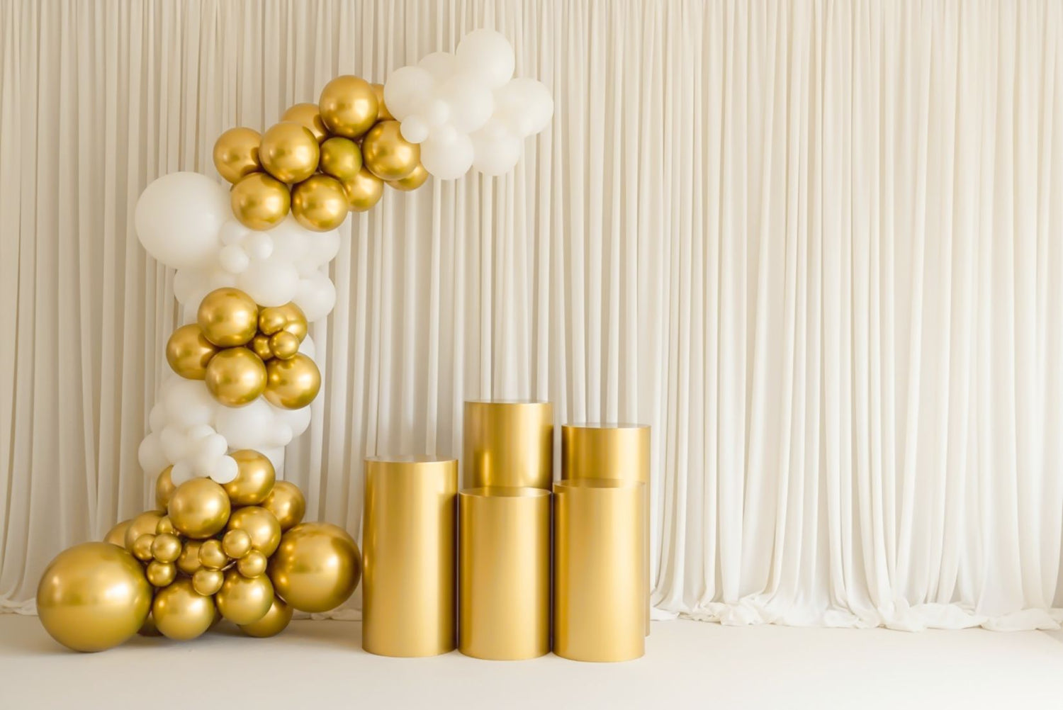 Decorative setup with gold balloons and five piece gold dessert cylinders against a white curtain background