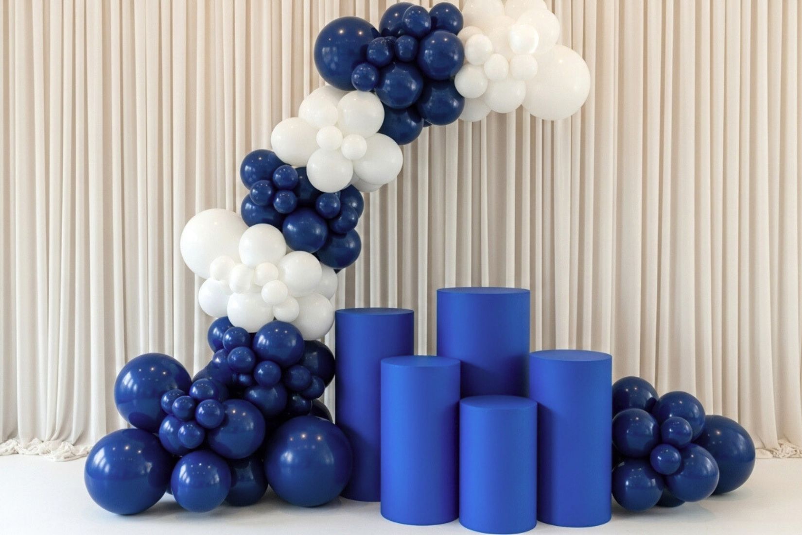 Decorative arch made of blue and white balloons with blue cake pedestal stands against a beige curtain background.