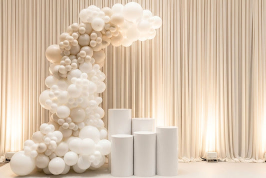 White balloon arch and five piece dessert cylinders for decorations against a beige curtain backdrop