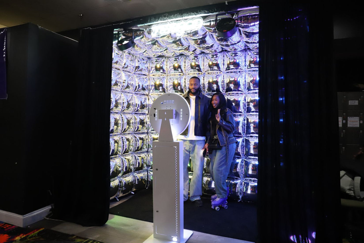 Two people standing inside an immersive cube with a large display of foil balloons and an open-air photo booth.