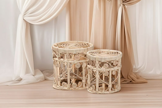 Two rattan wicker end tables in front of pipe and drape on a wooden floor with draped curtains in the background for a boho themed event.