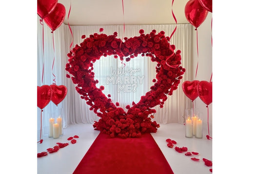 Heart-shaped floral arrangement with 'Will you marry me?' message, surrounded by red balloons and candles on a white floor.
