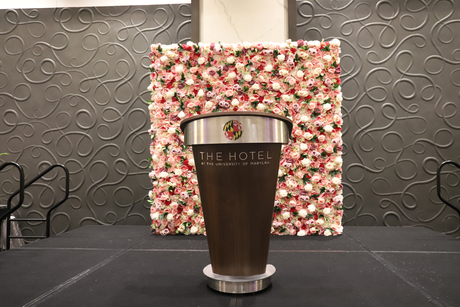 University of Maryland podium with "The Hotel" branding and a pink floral wall backdrop in front of a textured wall.