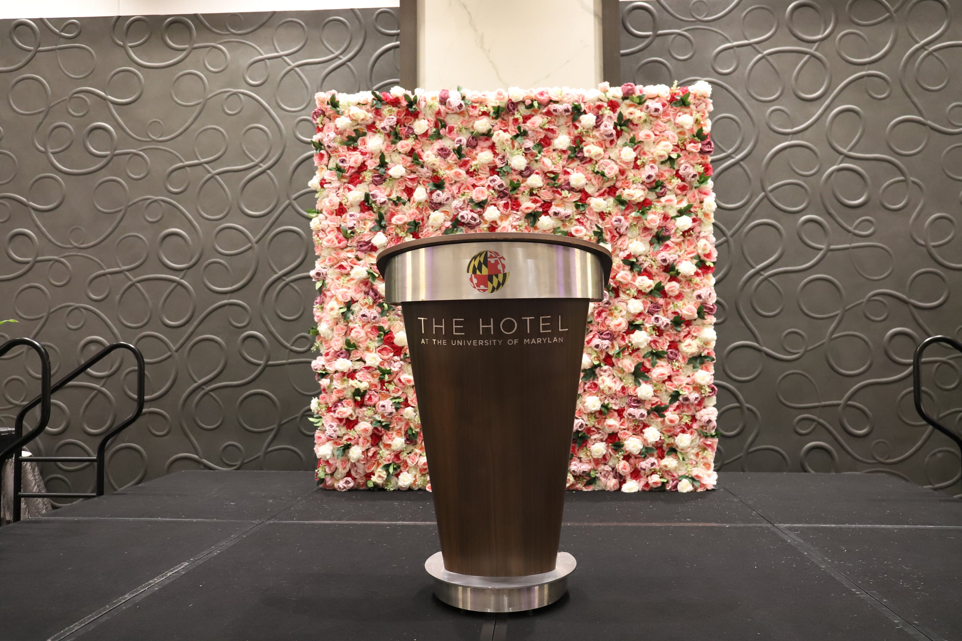 University of Maryland podium with "The Hotel" branding and a pink floral wall backdrop in front of a textured wall.