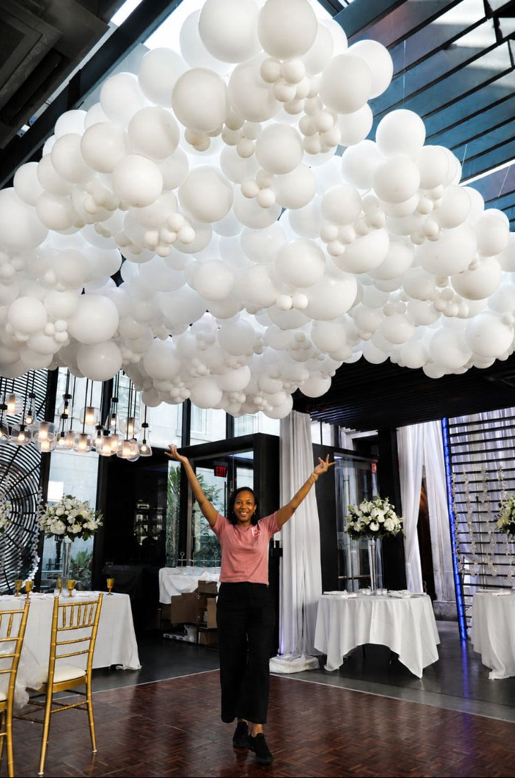 A woman stands with her arms raised under a large balloon cloud installation at the Dirty Habit, DC made of many white balloons, set in an indoor event space with tables and chairs.
