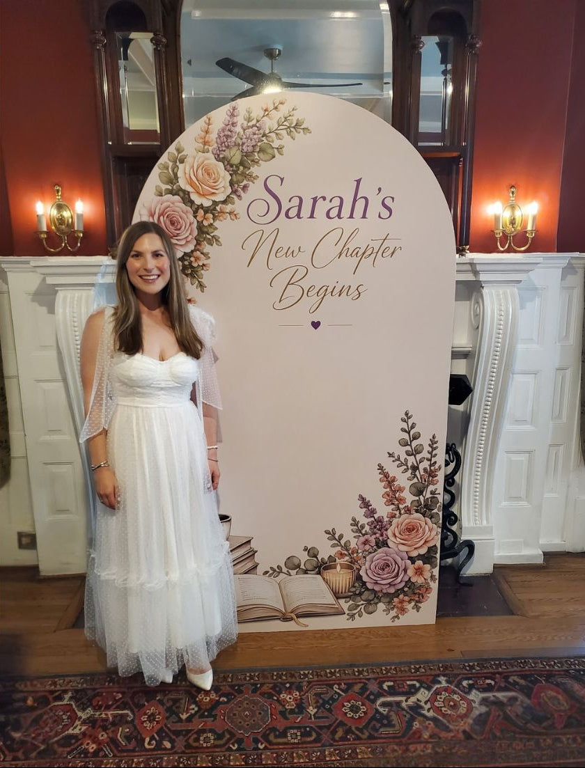 A woman in a white wedding dress stands beside a large personalized arch backdrop that reads 'Sarah's New Chapter Begins' with floral illustrations, set in an indoor event space with warm wall lighting and a patterned rug.