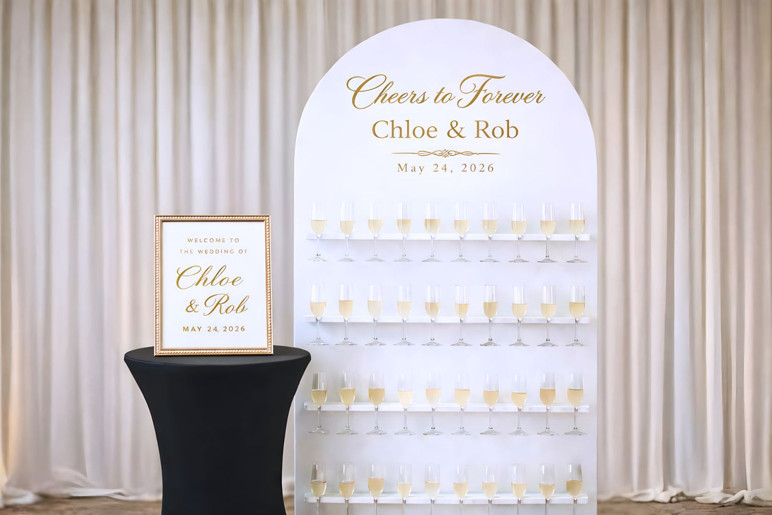 Decorative champagne wall arch with 'Cheers to Forever Chloe & Rob' text and wine glass illustrations, on a black stool against a white curtain background.