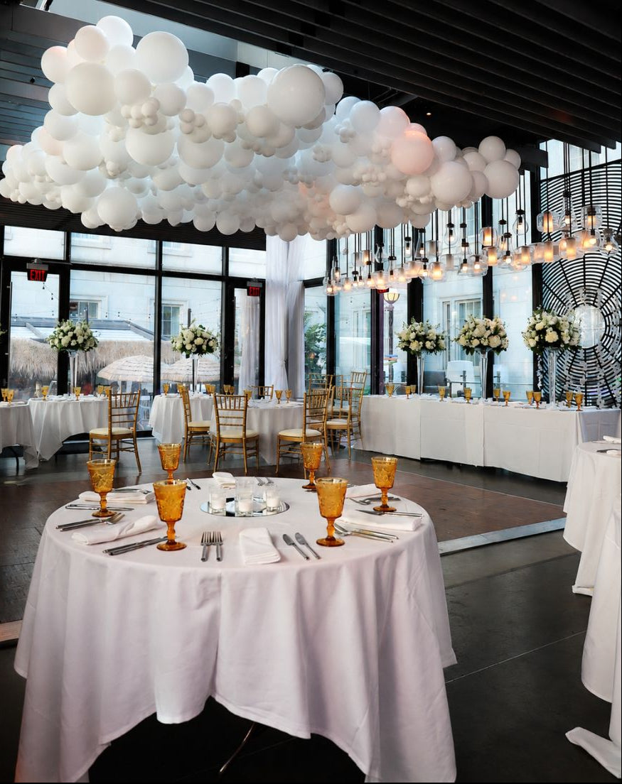 A decorated event space with round tables covered in white tablecloths, amber goblets, and silver flatware, under a large cloud-like balloon installation hanging from the ceiling.