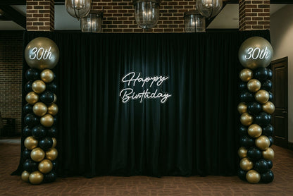 Decorative setup with balloons and a large 'Happy Birthday' neon sign on a black pipe and drape setup.