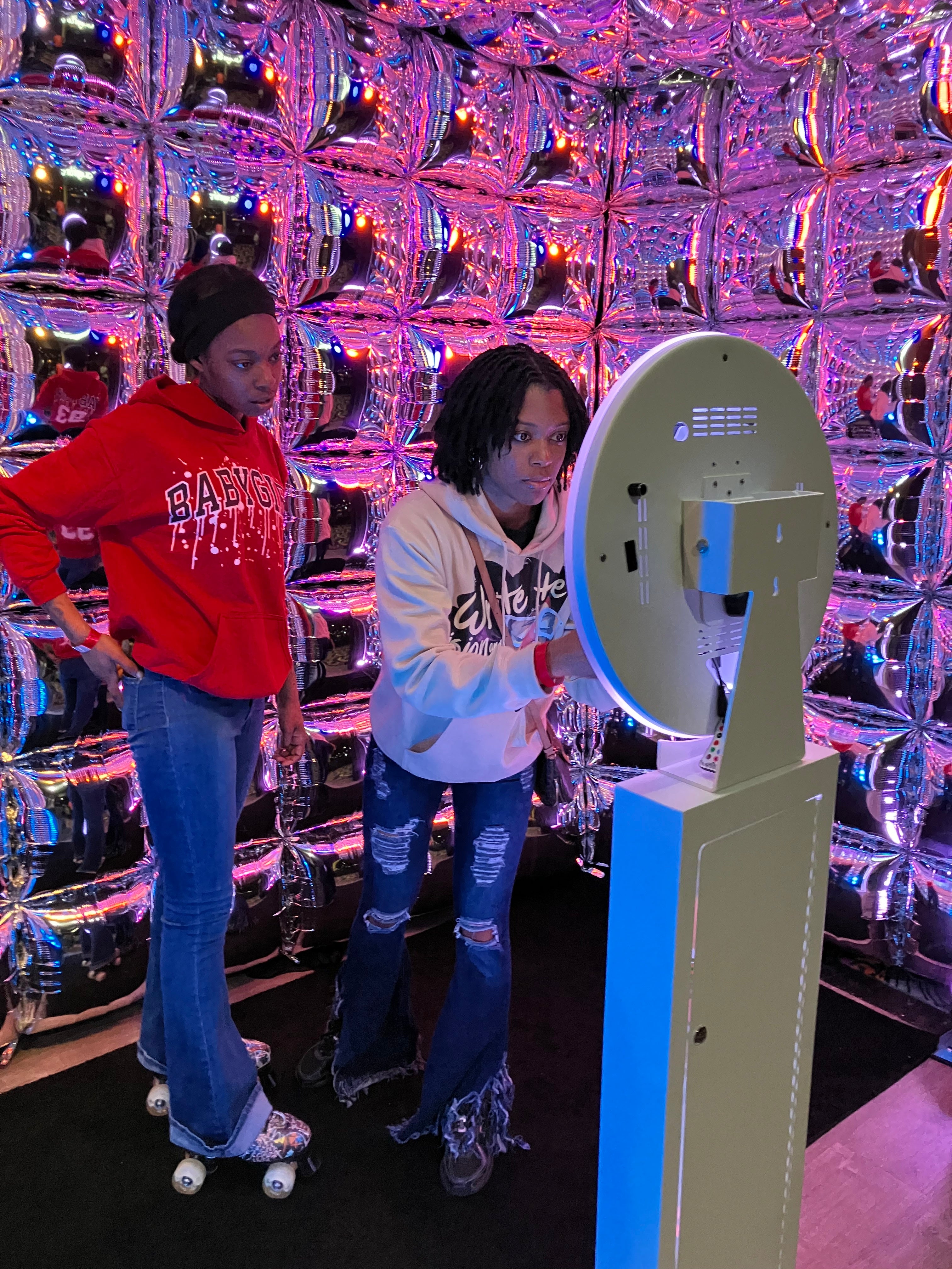 Two people standing in front of a mirrored balloon wall with a photo booth camera with LED neon lights.