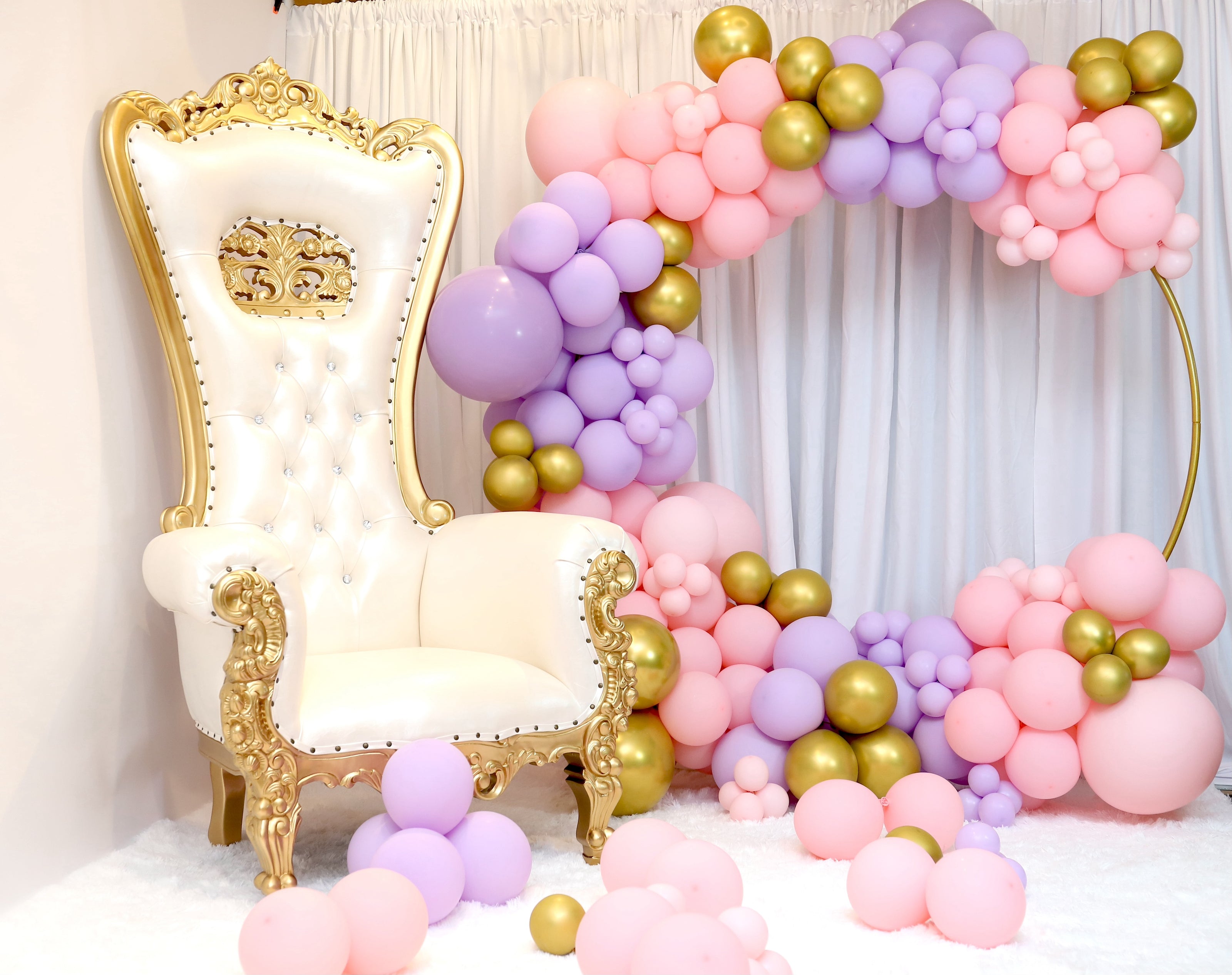 Decorative setup with a white throne chair, gold accents, and pink, purple, and gold balloons against a white curtain.