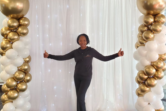 Person standing between two balloon columns with gold, white, and black balloons against a white curtain background.