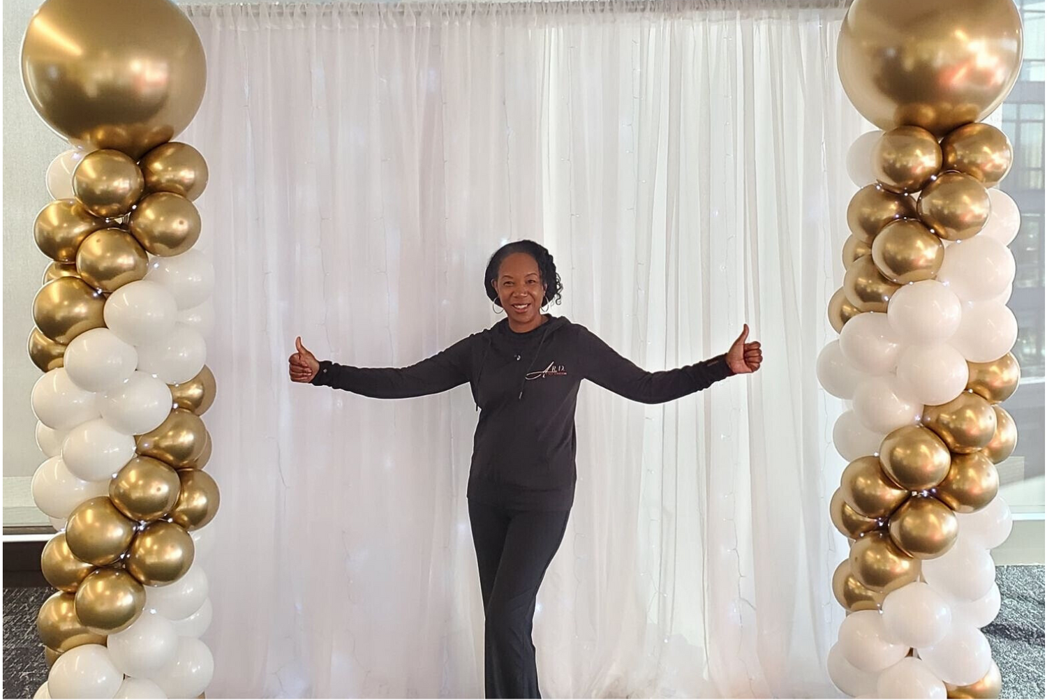 Person standing between two balloon columns with gold, white, and black balloons against a white curtain background.